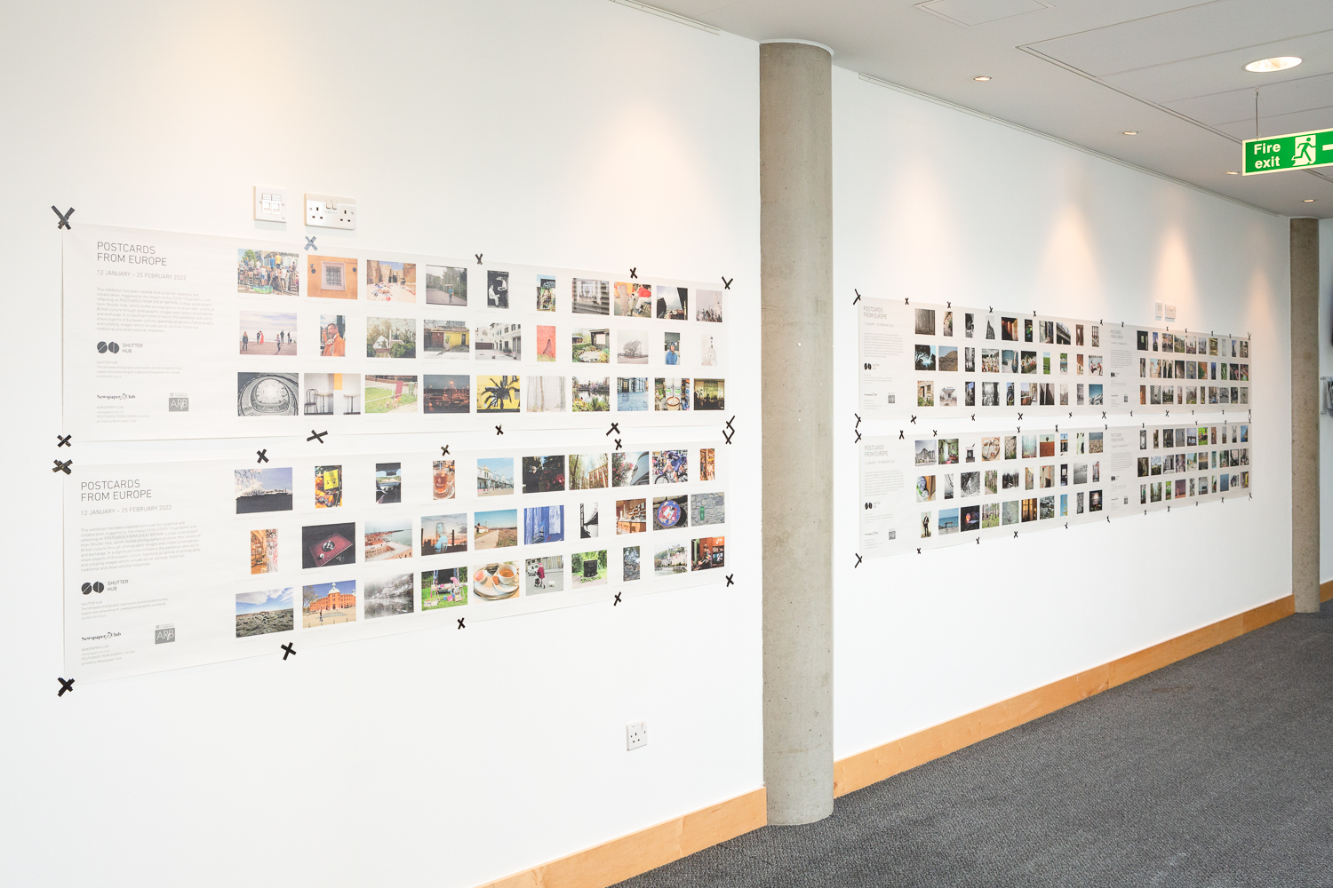 Installation photograph of 'Postcards from Europe' exhibition, showing postcard-sized photographs printed on long strips of newsprint, taped to a white wall, with concrete pillars between the sections.