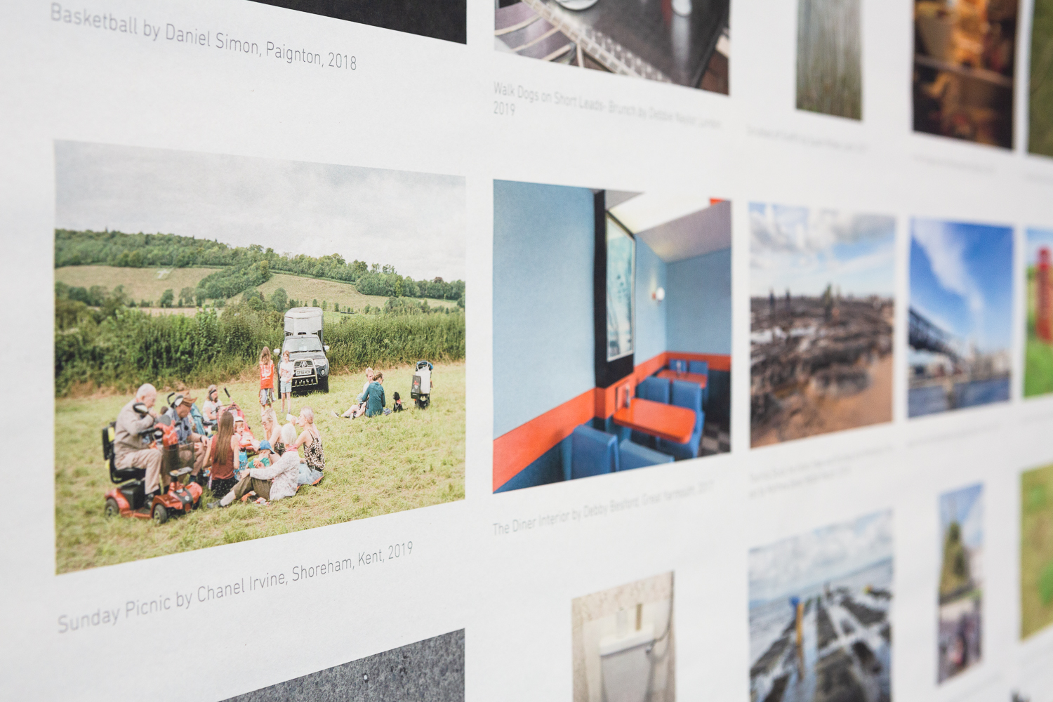 Installation photograph of 'Postcards from Europe' exhibition, showing a detail of postcard-sized photographs printed on long strips of newsprint, an image of a family on a picnic by a car in a field is visible in the foreground, with a photograph of an empty cafe to the centre of the image