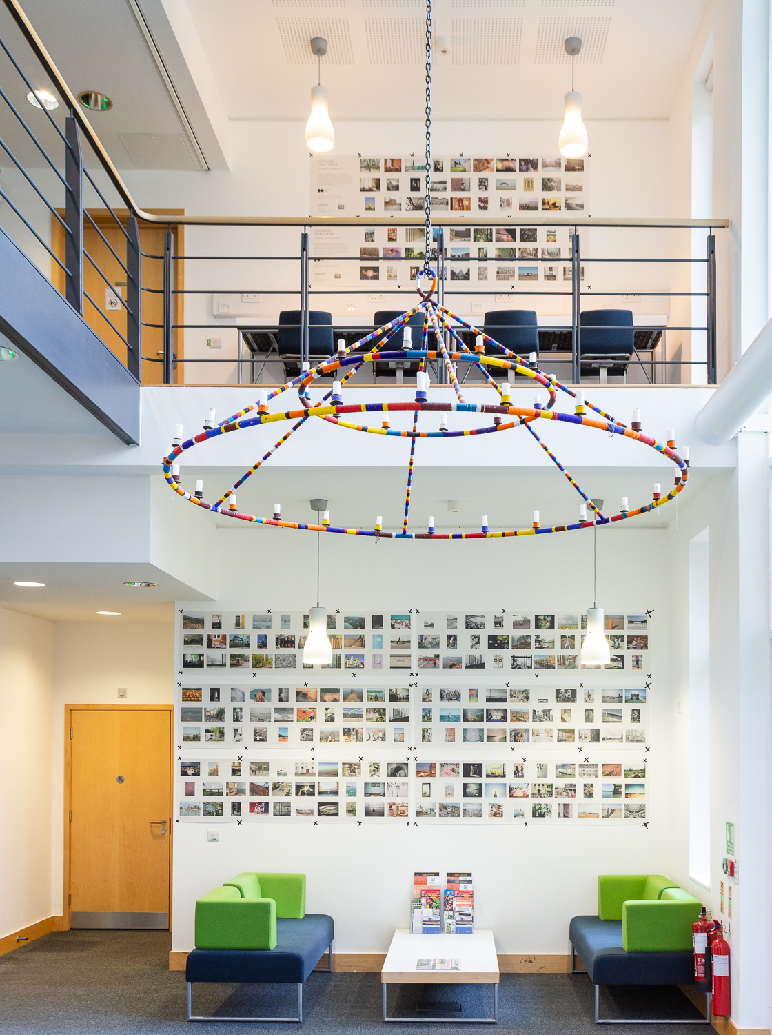 Installation photograph of 'Postcards from Europe' exhibition across two floors, showing postcard-sized photographs printed on long strips of newsprint, taped to the wall, with a lighting installation suspended from the ceiling in front, green and blue sofas and a table below and a wooden door to the left.