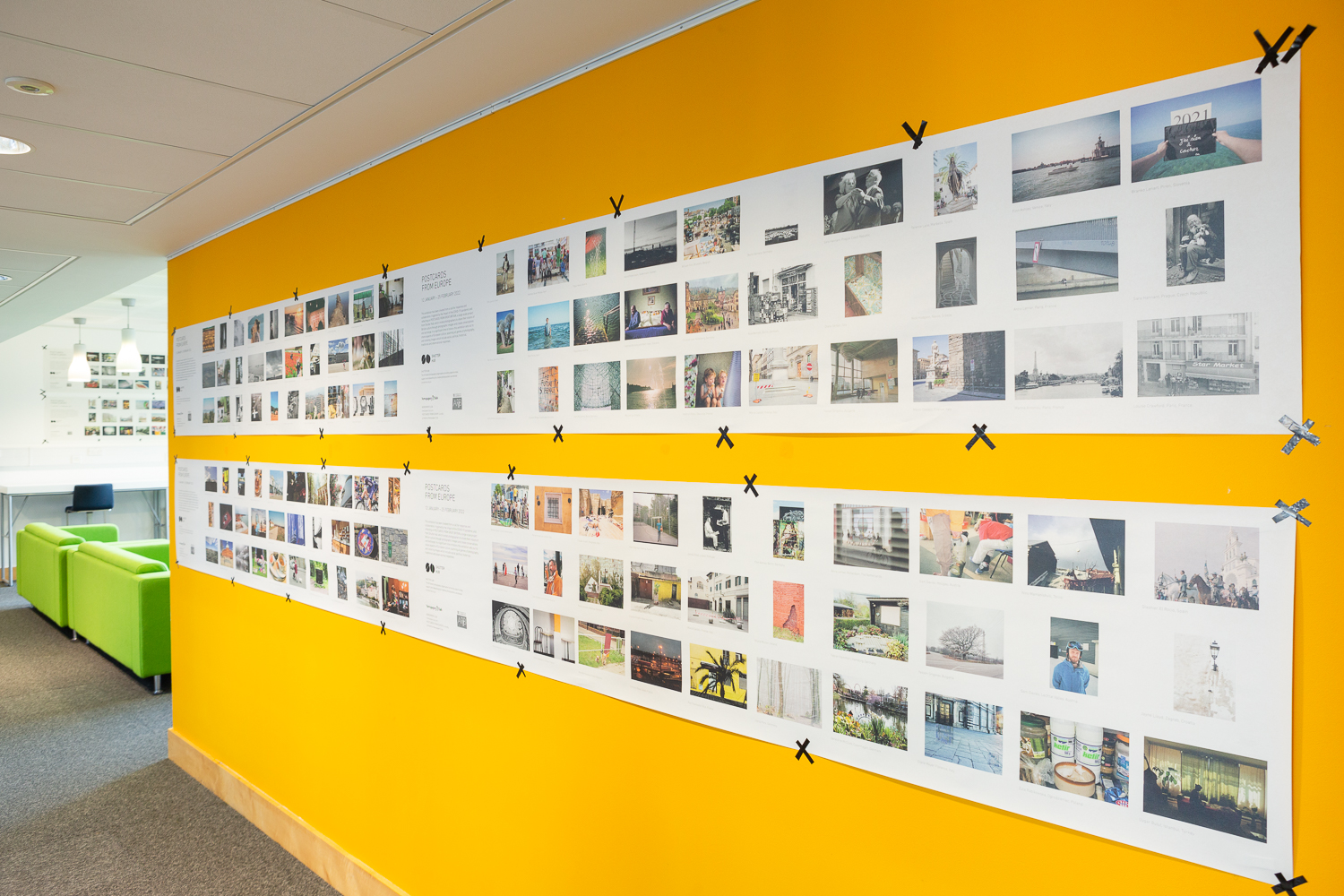 Installation photograph of 'Postcards from Europe' exhibition, showing postcard-sized photographs printed on long strips of newsprint, taped to a yellow wall, with green sofas from a seating area visible in the distance to the left of the frame.