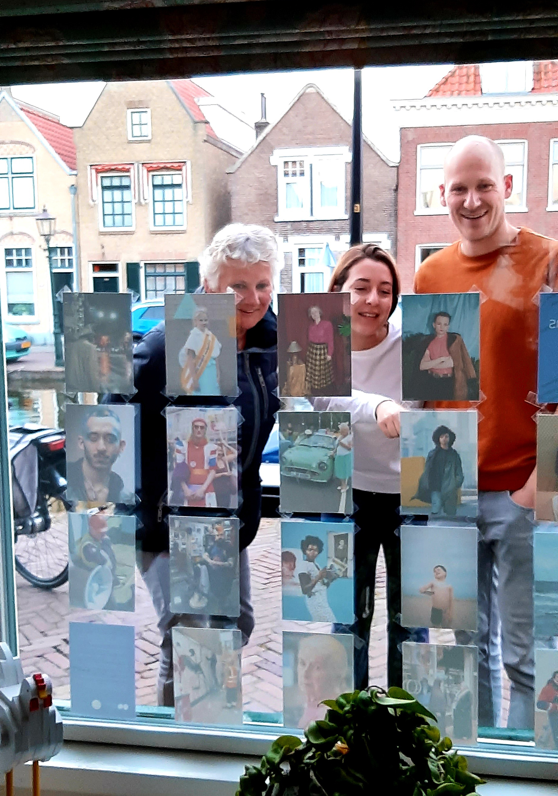 Photograph looking out of a window displaying postcard-sized photographs, with three people looking at the images and smiling, with buildings behind them in the background.