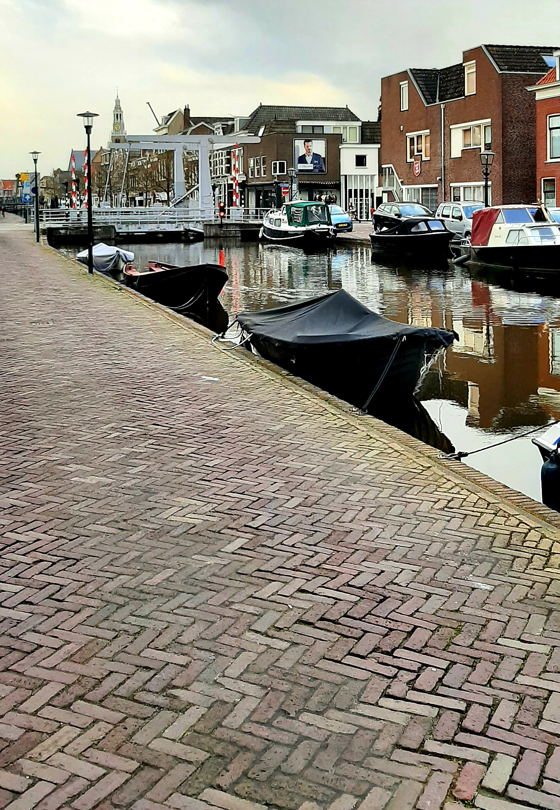 Photograph showing a canal with boats (some covered in protective fabric), buildings in the distance and a brick footpath in the foreground