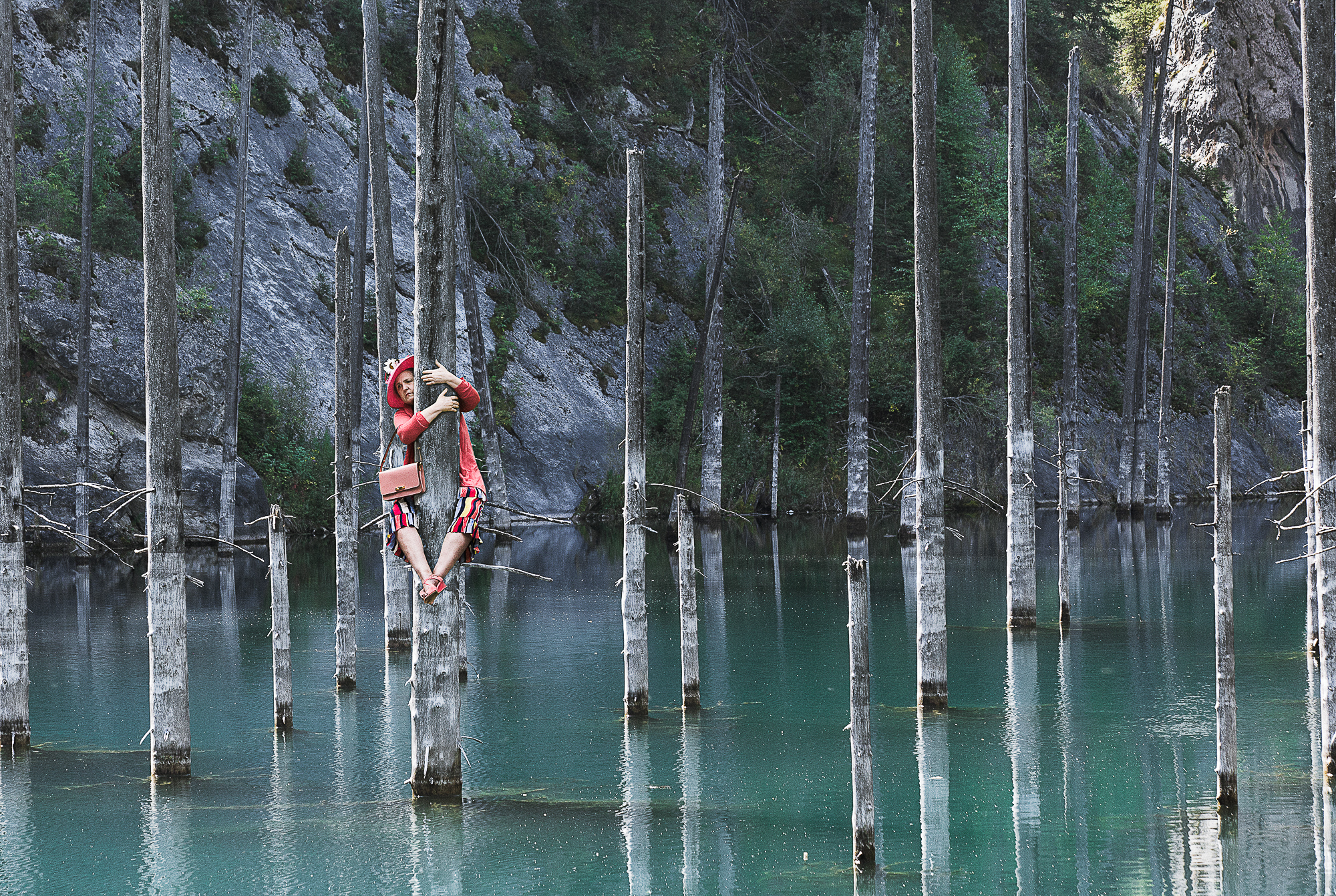 Photograph of a woman dressed for a garden party clings to a dead tree in the middle of a lake