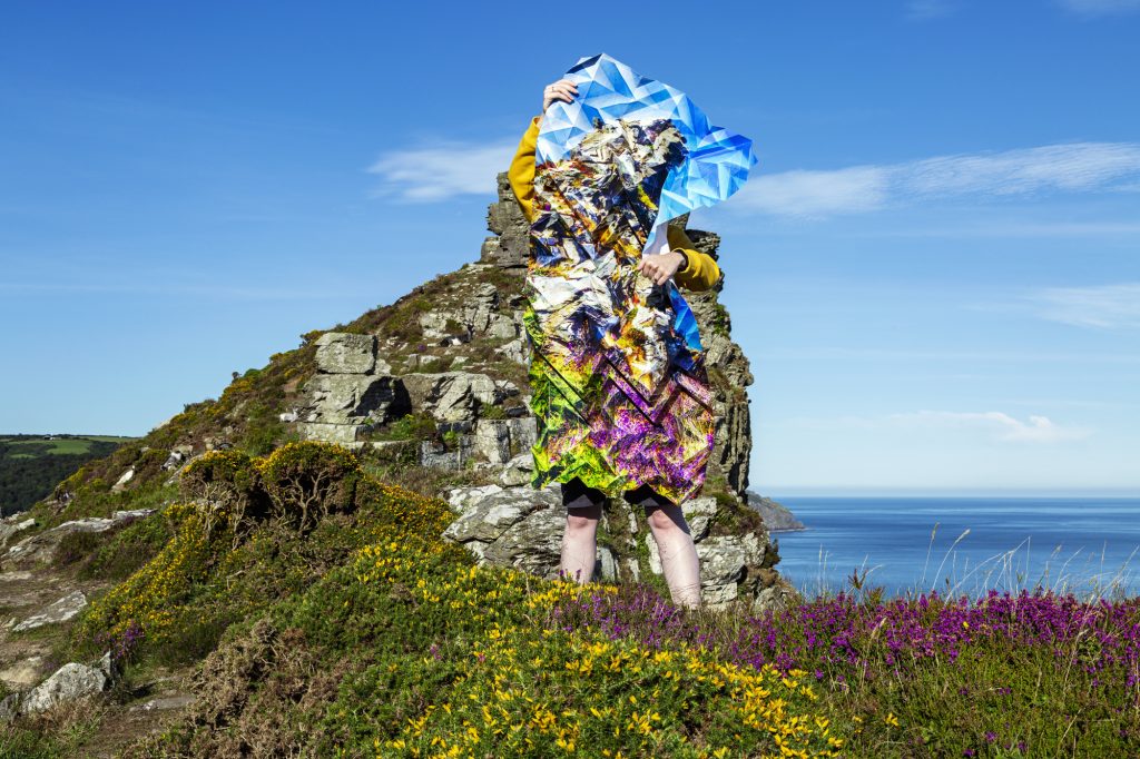 Image of a woman standing on a cliff top and holding a large, intricately-folded, photograph that blends in with the landscape.
