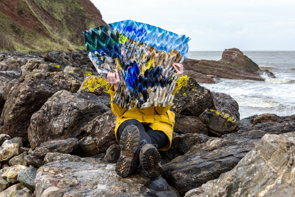 Image of a woman sitting on a rocky shoreline and holding a large, intricately-folded, photograph that blends in with the landscape.