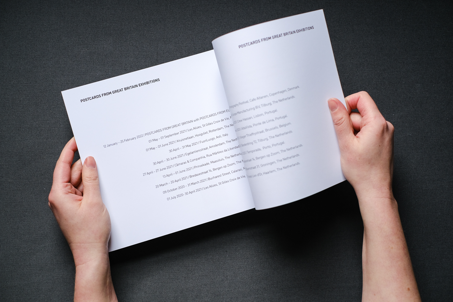 Photograph of hands holding the Postcards from Great Britain catalogue open on a double page spread with text listing exhibition locations, on a dark grey fabric background.