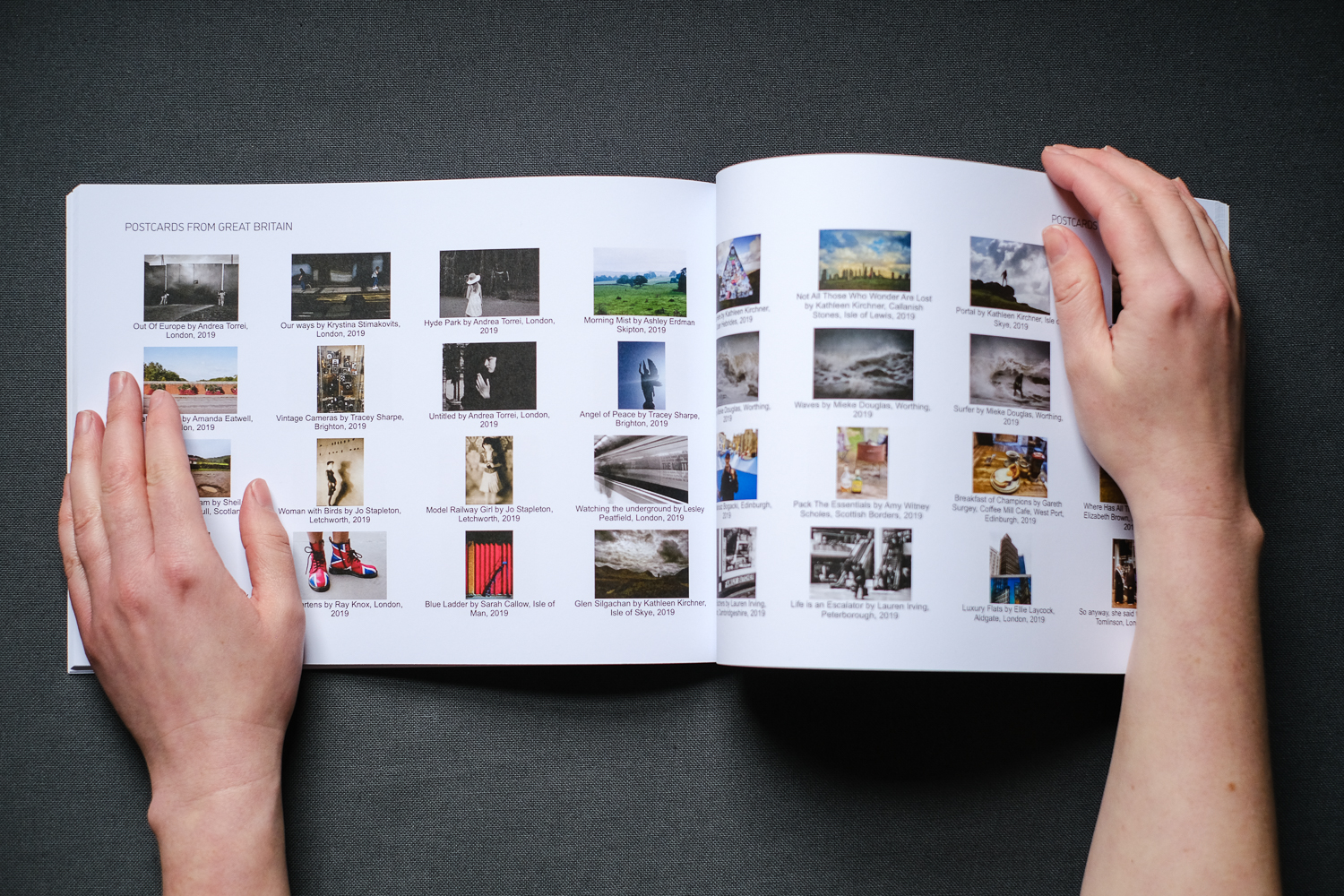 Photograph of hands holding the Postcards from Great Britain catalogue open at a double page spread, showing images from the archive across the pages and 'POSTCARDS FROM GREAT BRITAIN' in text at the top of each page, with a dark grey fabric background.