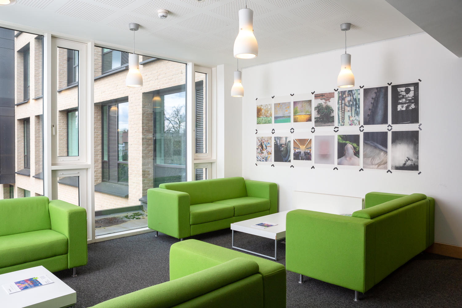 Photograph of Everything I Ever Learnt, a Shutter Hub exhibition with Art at the ARB at Cambridge University, showing a space with newsprint images taped to the wall on the left, green sofas in front, and external windows visible through the large window to the right.