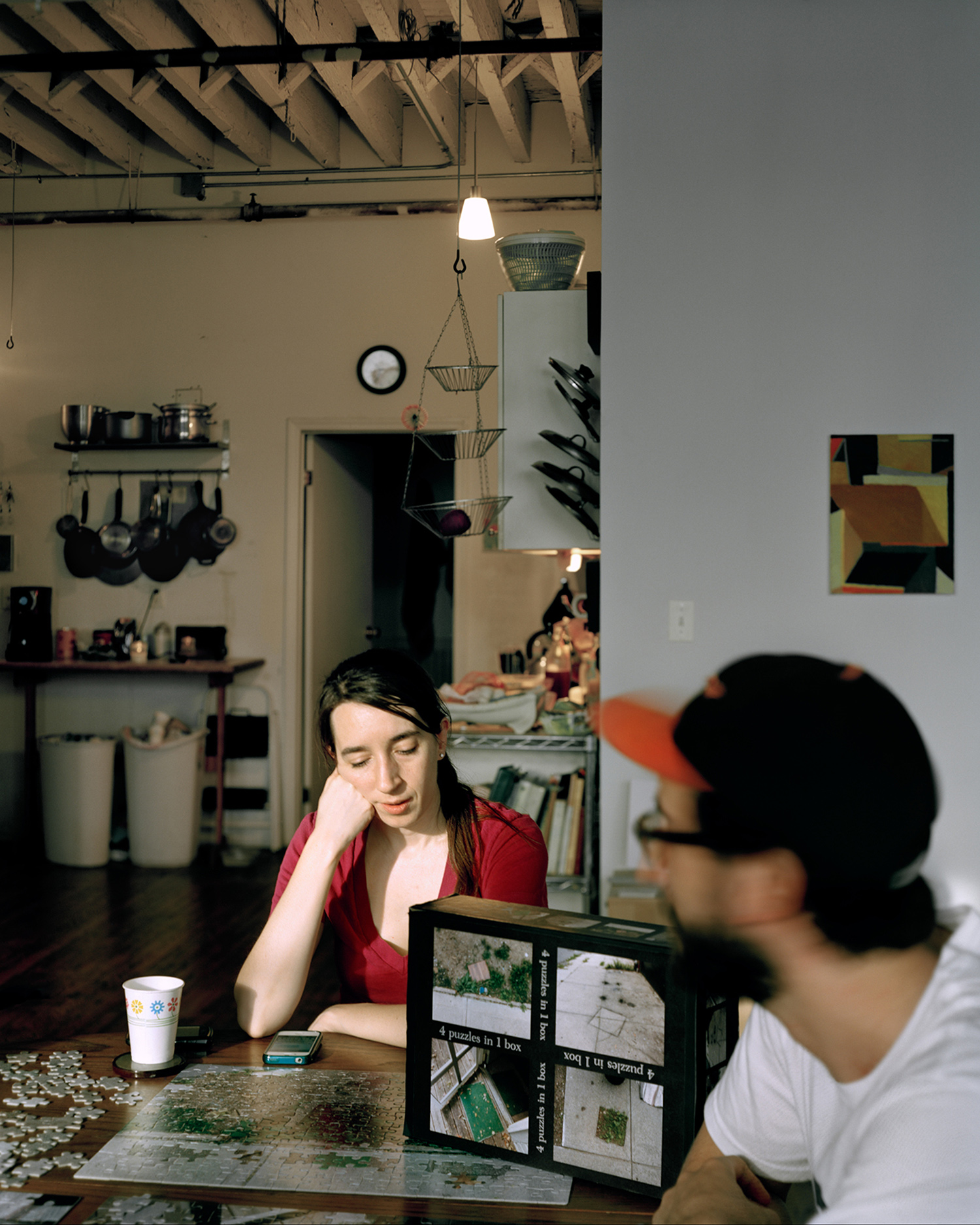 A woman and a man sit at a table playing with a jigsaw picturing urban scenes. A painting hangs behind them near the kitchen.