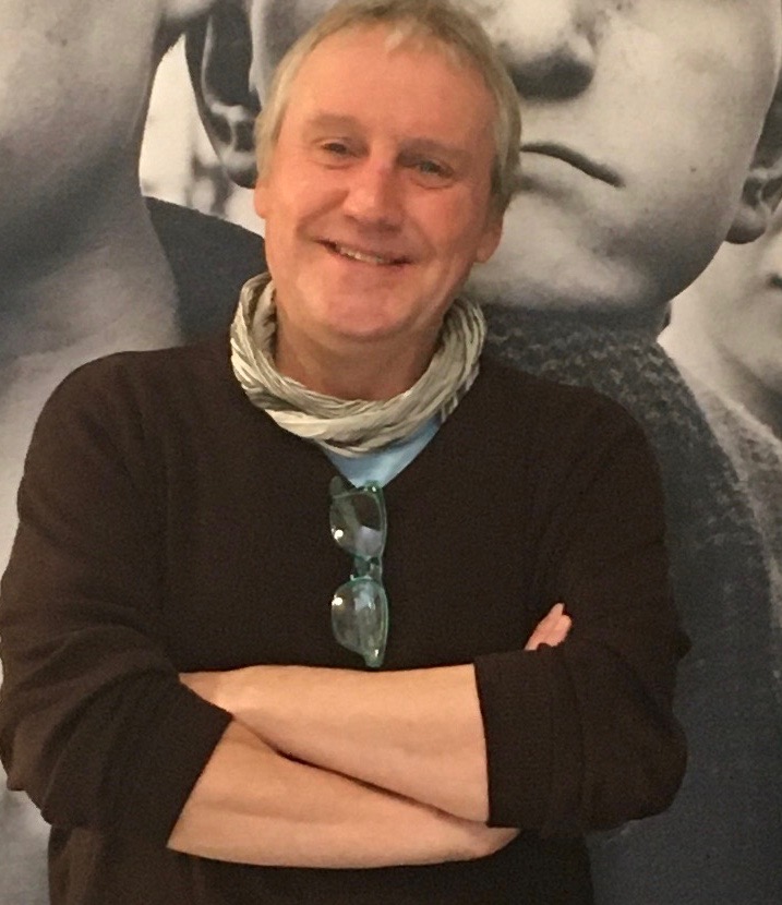 Photographic portrait of Malcolm Dickson, standing smiling with arms crossed in front of a large photographic print