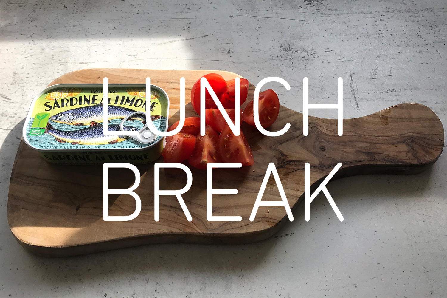 Photograph of a wooden board with a tin of fish and a cut tomato on a gray marble table with the words 'LUNCH BREAK' in white over the centre of the image