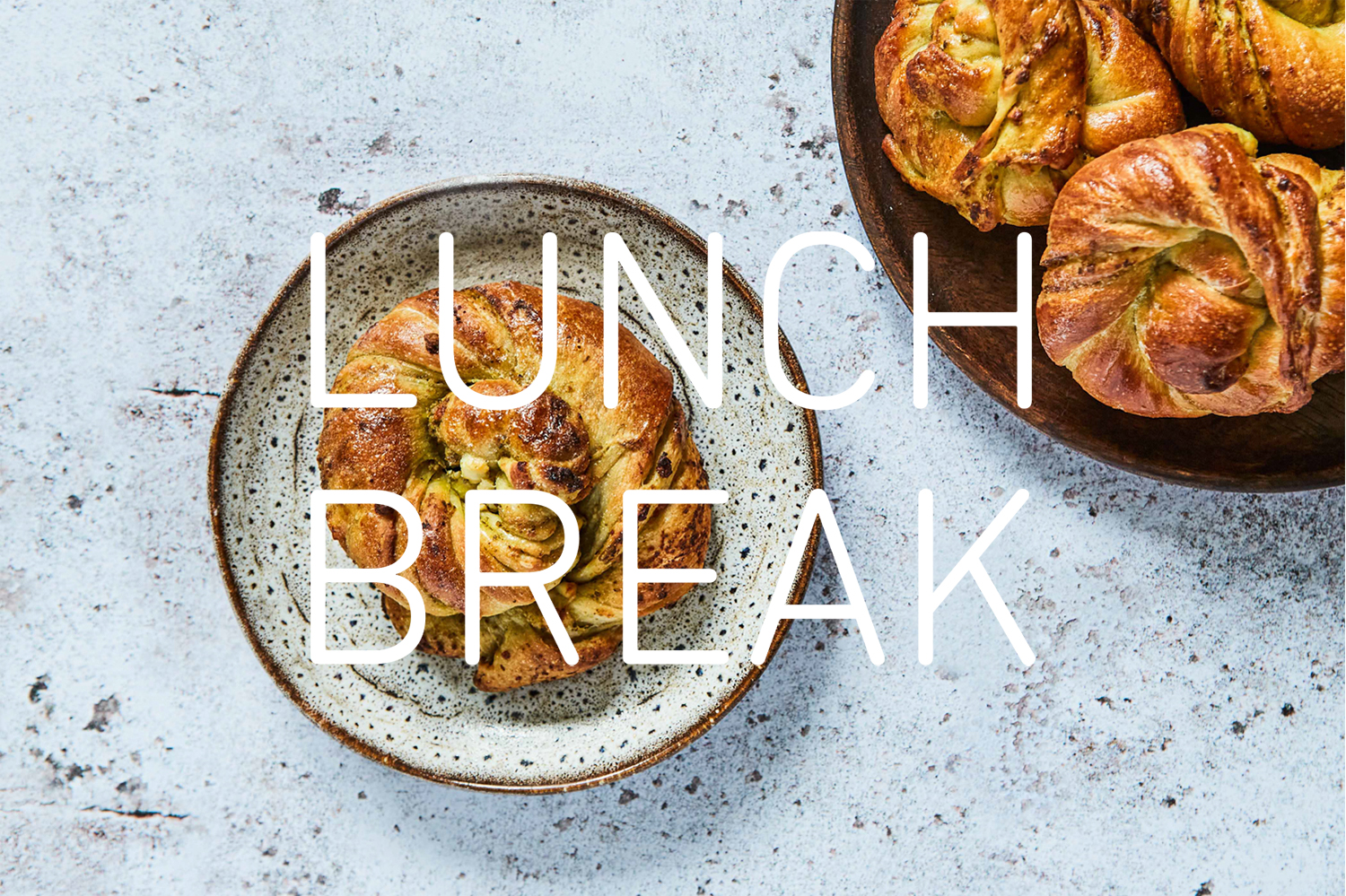 Photograph of a pastry on a dish on a weathered white table, with the words 'LUNCH BREAK' in white over the top