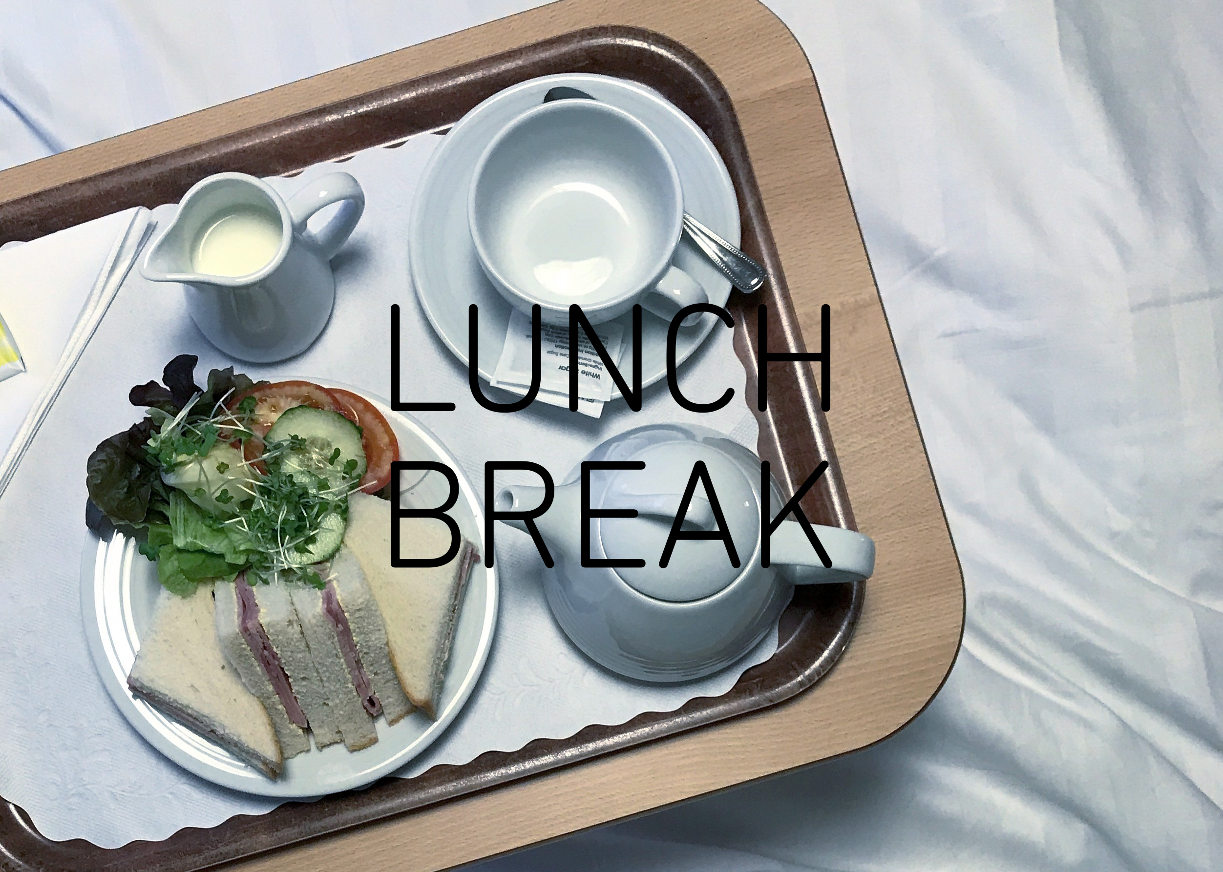 Photograph looking down on a tray with triangular ham sandwiches and a side salad, a small jug of milk, cup and saucer and small teapot, with the words 'LUNCH BREAK' in the centre