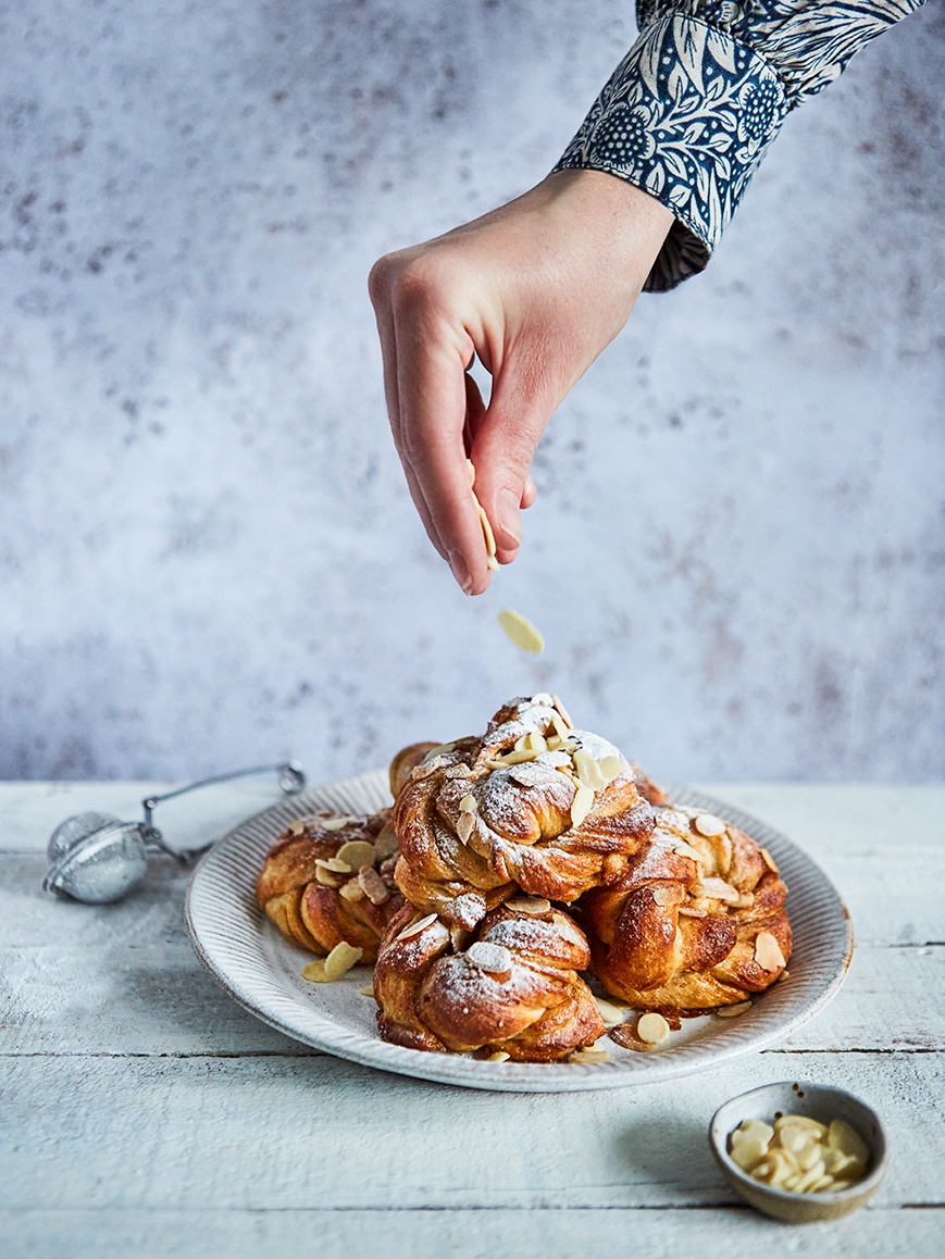 Swedish buns on a plate, with a hand sprinkling chopped almonds from above