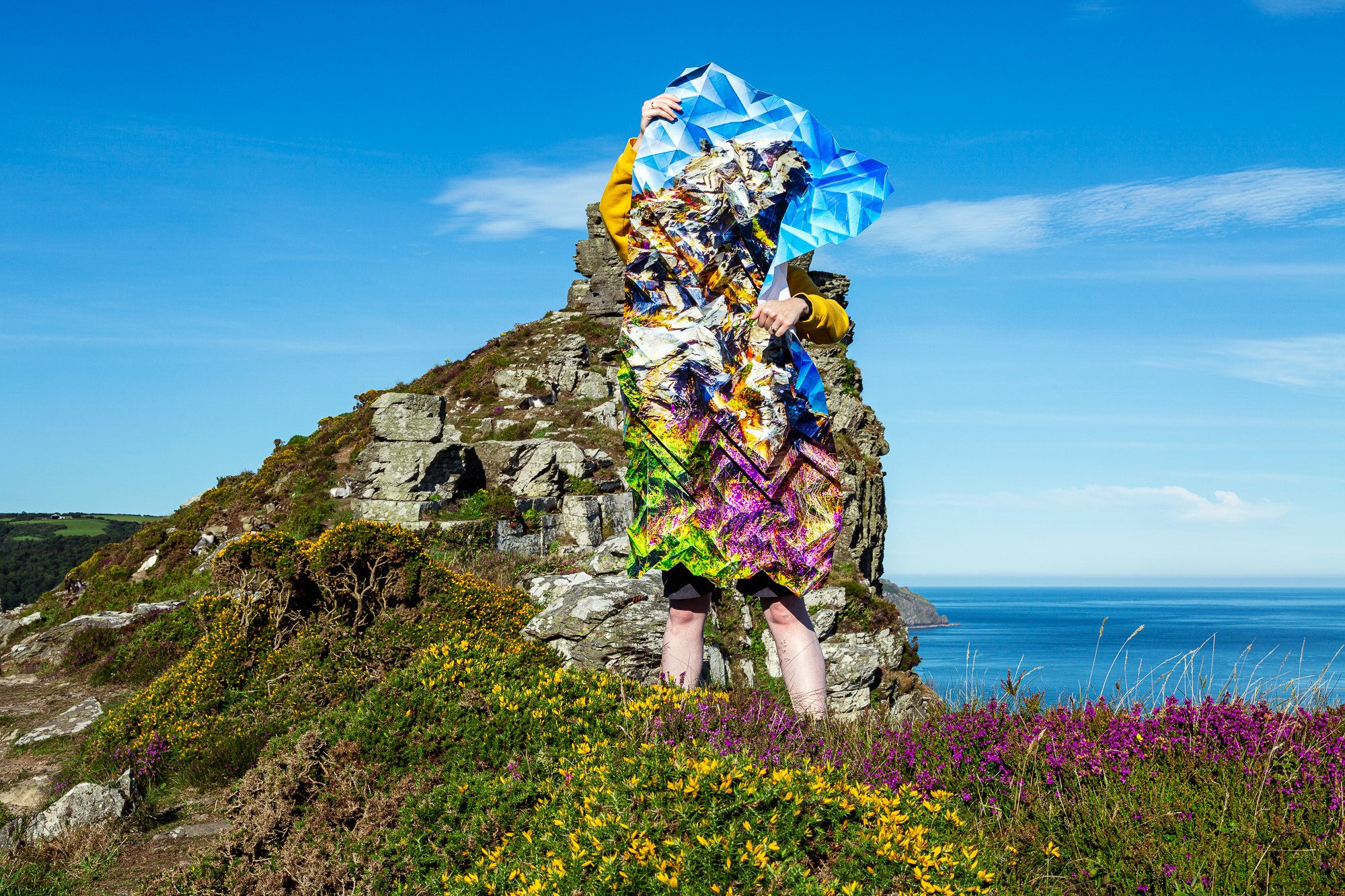 Image of a woman standing on a cliff top and holding a large, intricately-folded, photograph that blends in with the landscape.