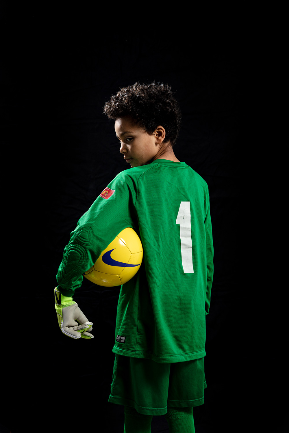 Child wearing a green goalkeeper's uniform with the number '1', looking over their shoulder, holding a yellow football under their arm