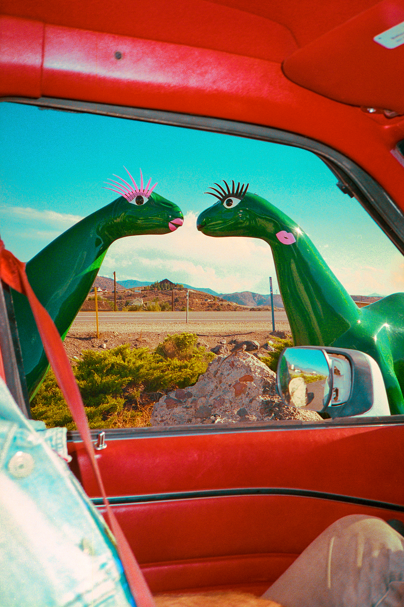 Dinosaur statues outside a gas station entrance, as a lizard looks up from the rock below. Taken through a driver's side window in September of 2020, during the coronavirus pandemic.