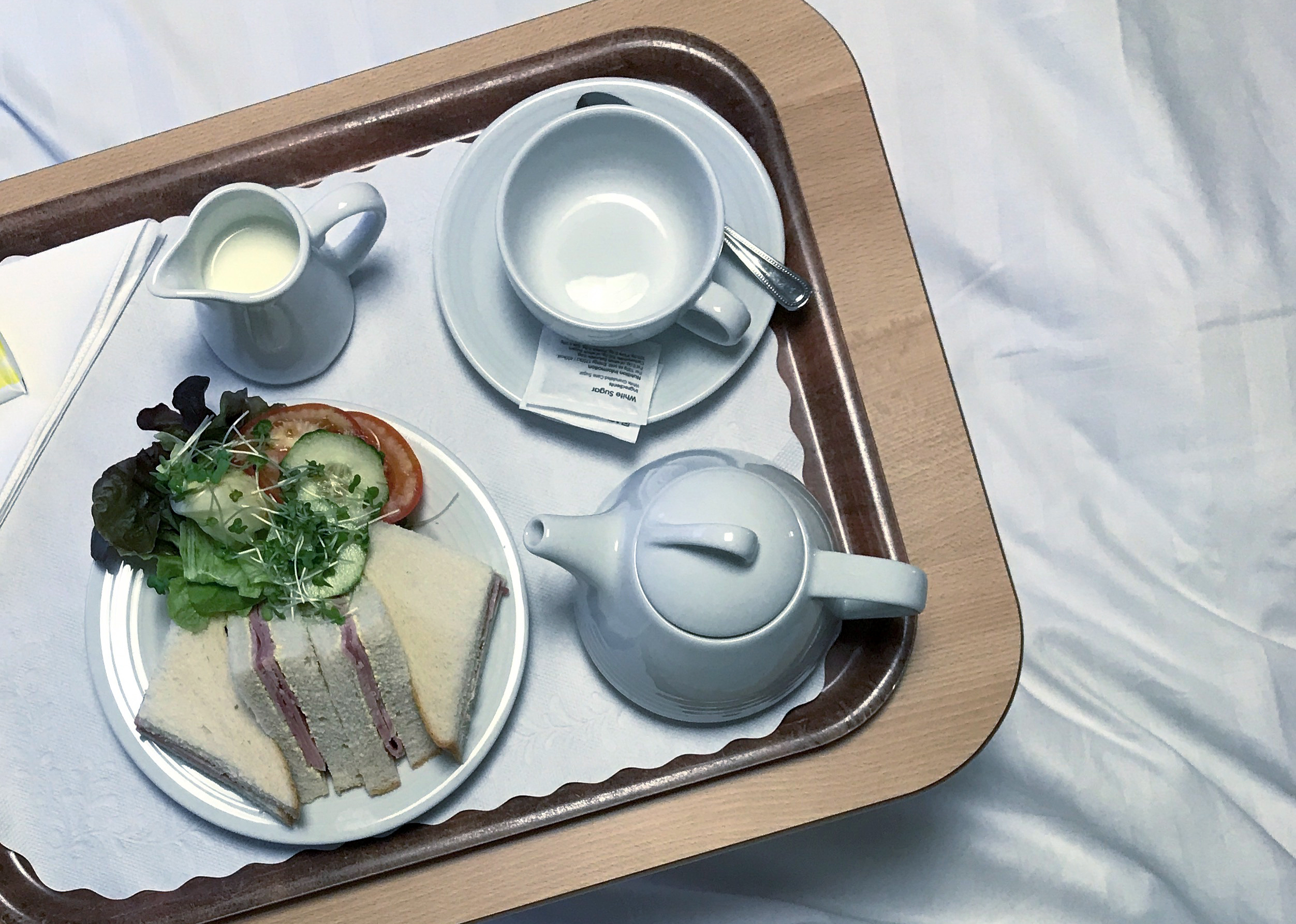 Photograph looking down at a tray containing an empty cup and saucer, a small teapot, a jug of milk and a plate with triangular ham sandwiches and salad to the left