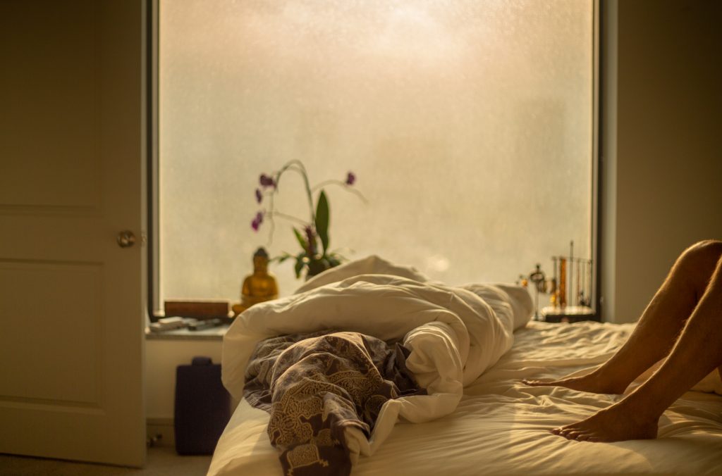 A man’s legs are seen in a rumpled unmade bed with sun pouring through a large window behind.