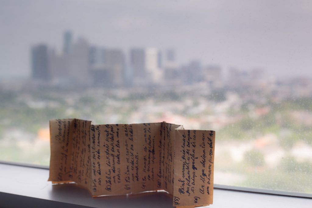 An old letter folded like a concertina, stands sideways on a window sill, with a downtown skyline seen beyond.