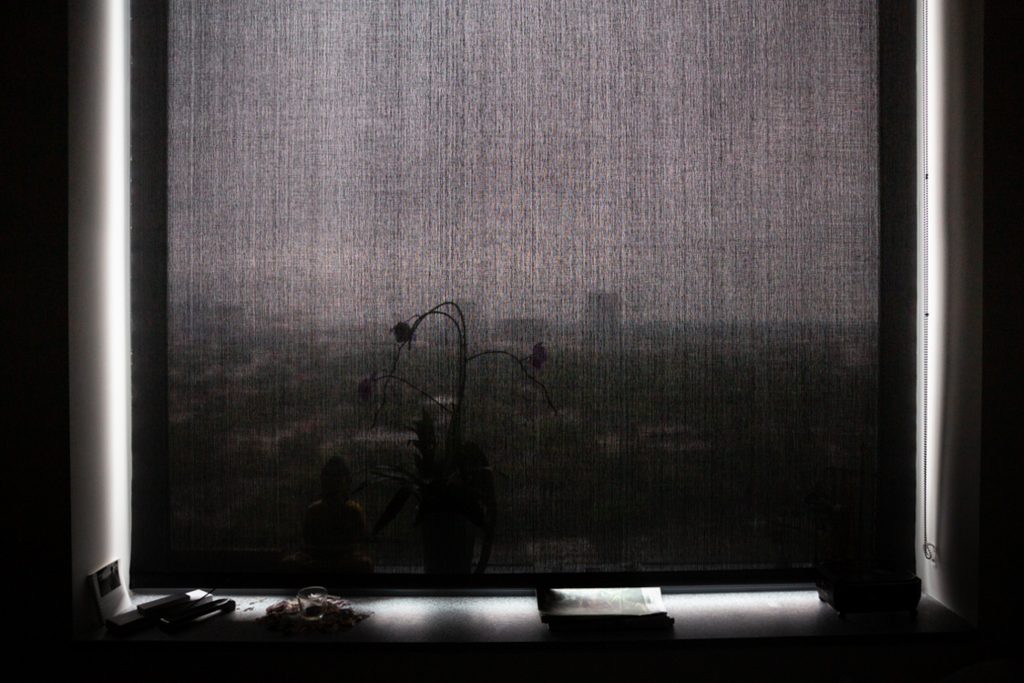 A black window blind is pulled down in front of a window with light coming in around the edges of it and the city view outside just seen through the semi opaque fabric.