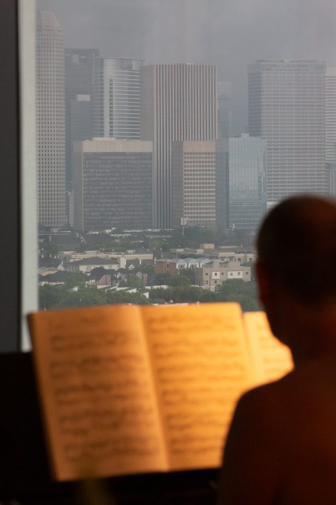 A man is silhouetted playing piano in front of a large window with a view of skyscrapers seen outside.
