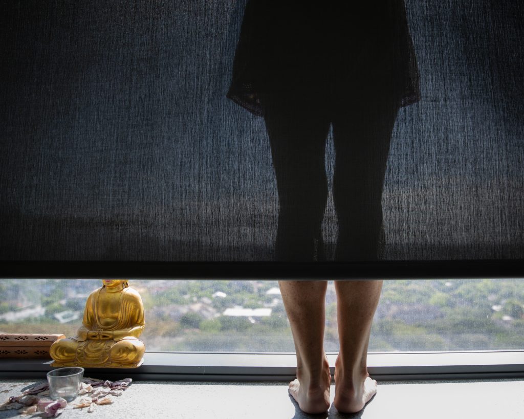 A woman’s legs are seen standing on a window sill peeping out from beneath a black blind pulled down below the knees, the figure seen silhouetted through the semi opaque fabric.