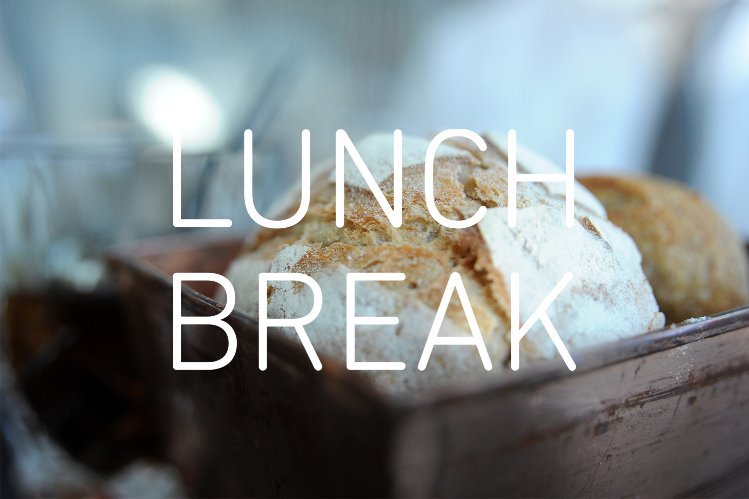 Photograph showing a loaf of break in a pan, with flour and the crust visible, selective focus so just the top is sharp. The words 'LUNCH BREAK' are overlaid in white in the centre.